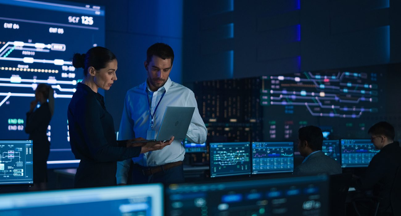 Project Leader and Chief Executive Discuss Data Shown on a Laptop. In Background Big Screen Showing Infographics of Infrastructure, Charts, System Analysis. Telecommunications Control Room